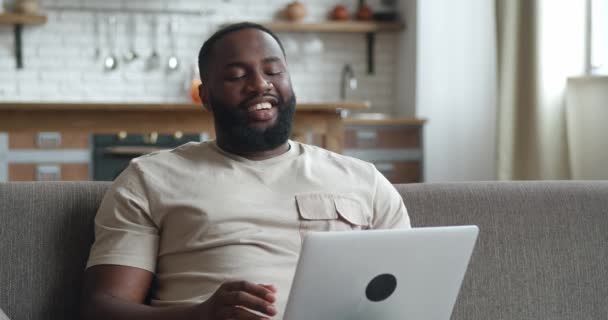 自宅でソファの上でビデオ会議でパートナーと話しているビジネスマン。男性のフリーランスは、ビデオ通話でオンライン会議のためのラップトップコンピュータを使用しています。家庭から働き、遠隔で働き、社会的孤立. — ストック動画
