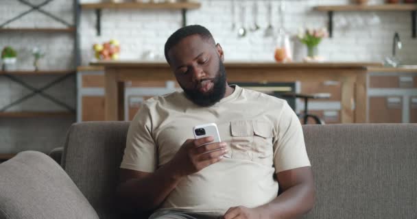 自宅でソファに座っているスマートフォンを使用して黒人男性に乗り込み、疲れている若いアフリカ人男性は、画面を見て携帯電話を保持しています読書電子書籍アプリオンライン音楽を聞く — ストック動画