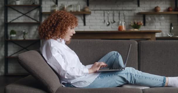 Sidovy av upphetsad rödhårig ung kvinna sitter på soffan på bärbar dator som arbetar i vardagsrummet. Hon får goda nyheter och är glad. Visar ja gest — Stockvideo