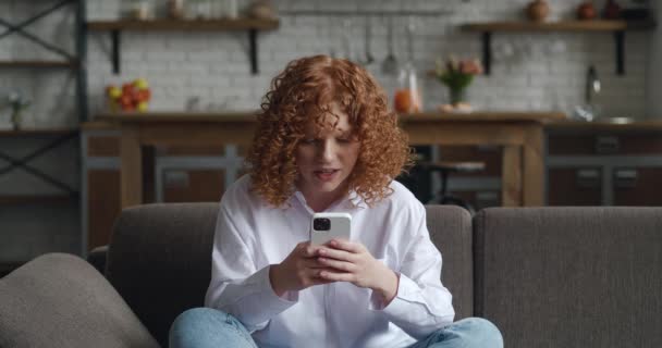 自宅のソファに座っているソーシャルメディアアプリでスマートフォンの勝利の贈り物や賞を使用して喜びの若い赤毛の女性。リビングルームでモバイル勝利を祝う興奮した幸せな女性の勝者,はいジェスチャーを示します — ストック動画