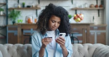 Oturma odasındaki koltukta oturan mutlu Afrikalı Amerikalı genç kadın, elinde kredi kartı ve cep telefonu ile online alışveriş yapıyor, dilek tutulan şeyleri alıyor, ev konseptinden alışveriş yapıyor.