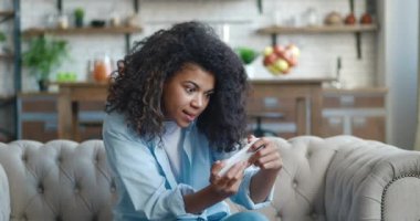 Genç Afro-Amerikan kadın akıllı telefonuyla heyecan verici mobil uygulaması ile oynuyor. Gülümseyen kadın, evdeki oturma odasında kanepede otururken yumruğunu sıkarak bir yarış oyunu kazanıyor..