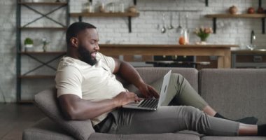 Gülümseyen Afrikalı adam kanepede dizüstü bilgisayar kullanarak oturma odasında yazı yazarken, genç işadamı bilgisayarda sohbet ederken internetten sosyal medya okurken, evde online çalışırken