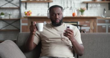 Akıllı telefon kullanan, kulaklıklı, gülümseyen siyah bir adam evdeki koltukta oturuyor, cep telefonuyla kahve içen Afrikalı genç adam ekrandan e-kitap uygulaması okuyarak internetten müzik dinliyor.