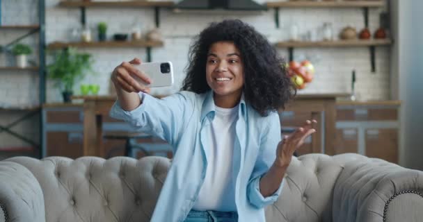 Sorrindo jovem afro-americana com cabelo encaracolado fazendo videochamada enquanto estava sentado em um sofá em casa. Retrato de mulher bonita fazendo um bate-papo por vídeo no fundo da sala de estar — Vídeo de Stock