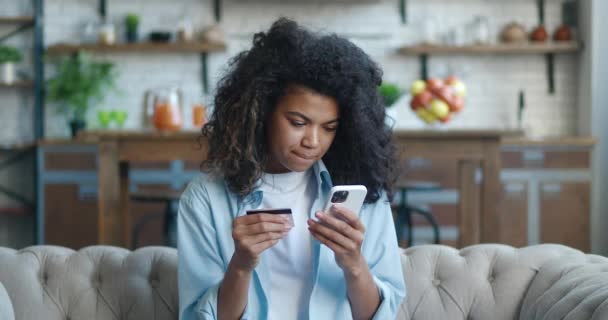 Feliz joven afroamericana sentada en el sofá en la sala de estar, con tarjeta de crédito y teléfono móvil para realizar transacciones en línea, comprar artículos deseados, disfrutar de compras desde el concepto de casa — Vídeos de Stock