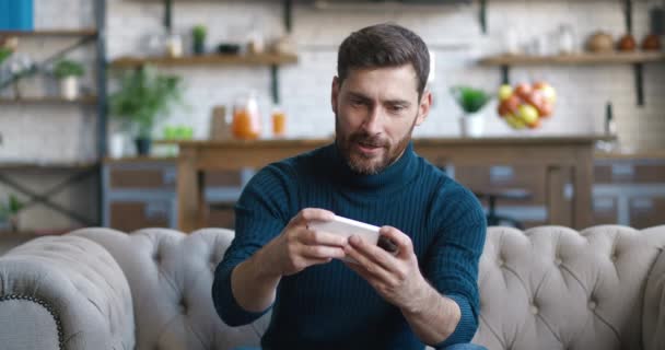 Szczęśliwy biały człowiek gracz grać w gry wideo online z aplikacji smartfon, siedzi na kanapie w domu, świętować zwycięstwo w grze, tak gest — Wideo stockowe