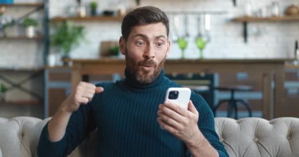 Verbazingwekkende gelukkige zakenman ontvangt bericht dat goed nieuws leest in de woonkamer. Opgewonden dolblij mannelijke winnaar vieren succes te kijken naar de smartphone zittend op de bank thuis. Mobiel overwinningsconcept — Stockvideo