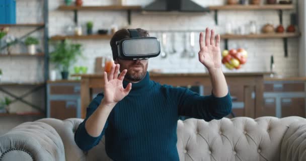 Ung man använder virtuella verklighets glasögon medan stiitng på soffan hemma. Millenial hane få erfarenhet av att använda VR-headset utforska cyberrymden i virtuella världar medan du sitter i vardagsrummet — Stockvideo