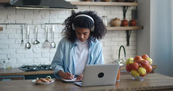 Afroamerikanische Studentin schreibt Notizen vom Laptop zu Hause. Mädchen im Kopfhörer, in der Küche sitzend. Akademikerinnen studieren konzentriert online. Bildungskonzept — Stockvideo