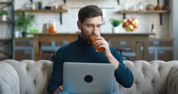 Joven hombre milenario freelancer utilizando el dispositivo portátil y beber jugo mientras está sentado en el sofá en la oficina en casa. Retrato de un hombre alegre trabajando a distancia en el ordenador en el sofá en la sala de estar — Vídeo de stock