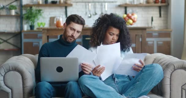 Rušný mladý pár drží čtení dokumentů papíry a při pohledu na notebook, zatímco pracuje z domova. Rodina sedí na pohovce kontrolní služba platit papírové domácí účty on-line na počítači diskutovat o rozpočtu. — Stock video