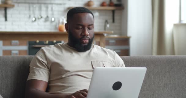 Joven afroamericano freelancer hombre usando el dispositivo portátil sentado en el sofá en la oficina en casa, retrato de un hombre trabajando a distancia en la computadora en el apartamento — Vídeo de stock