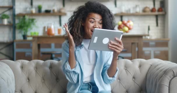 Ενθουσιασμένη νεαρή Αφροαμερικανή γυναίκα που χρησιμοποιεί tablet PC κερδίζοντας δώρο ή βραβείο στα μέσα κοινωνικής δικτύωσης app καθίσει στον καναπέ στο σπίτι. Ευτυχισμένη γυναίκα νικητής γιορτάζει κινητή νίκη στο σαλόνι, δείχνει ναι χειρονομία — Αρχείο Βίντεο