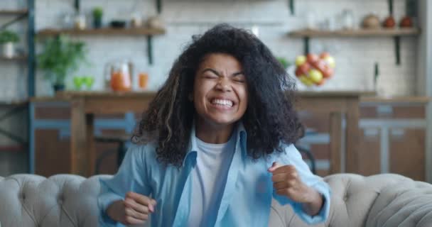 Kıvırcık saçlı genç Afro-Amerikan kadın TV 'de spor programı seyrediyor ve evde otururken evet işareti yapıyor. Genç dişiler zaferi kutluyor. Piyango kazanma konsepti. — Stok video