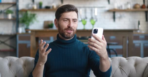 リビングルームでスマートフォンを呼び出し、携帯電話の画面上の人々に手を振って顔時間ビデオ通話を行う男の肖像画。若い男性ブロガーは、携帯電話を使用してvlogビデオブログを作る。オンラインコミュニケーションの概念 — ストック動画