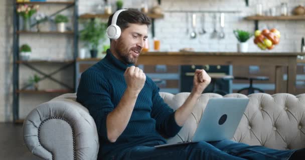 面白い幸せな若いビジネスマンは、ソファに座って自宅でラップトップで作業しながら、楽しいリスニング音楽を持つ無線ヘッドフォンを身に着けています。コンピューターを使った陽気なフリーランスはリビングルームで楽しむ — ストック動画