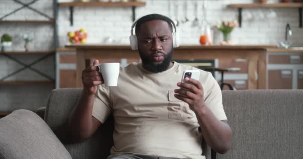 Sorrindo homem negro em fones de ouvido usando smartphone sentar no sofá em casa, jovem africano bebendo café segurando telefone celular olhando para a tela lendo e-book app ouvindo música online — Vídeo de Stock