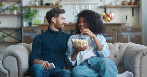 Alegre pareja multirracial ver la televisión y discutir viendo videos mientras come palomitas de maíz mientras está sentado en el sofá en casa. — Vídeos de Stock