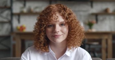 Kıvırcık saçlı neşeli kızıl kız oturma odasında kameraya bakıyor. Evde tek başına poz veren gülümseyen öğrenci kadın. İçerideki kendine güvenen kızıl saçlı genç bayanın vesikalığı.