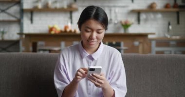 Mutlu Asyalı kadın, evde kanepede oturmuş akıllı telefon kullanıyor ve oturma odasında gülümsüyor. Evdeki koltukta otururken cep telefonuna ya da cep telefonuna mesaj bırakan bir kadın..