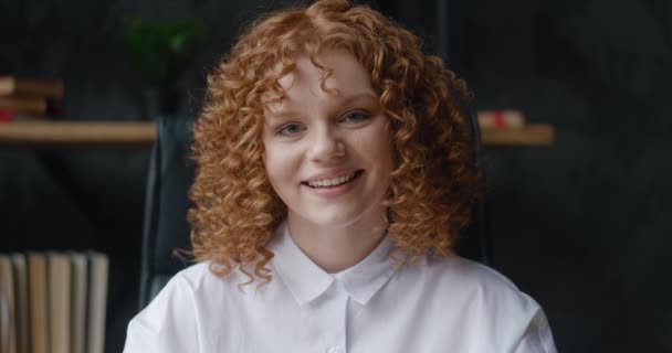 Крупним планом портрет усміхненої червонуватої молодої жінки студентки з кучерявим волоссям дивиться на камеру і мигтить її очі, на офісному фоні — стокове відео
