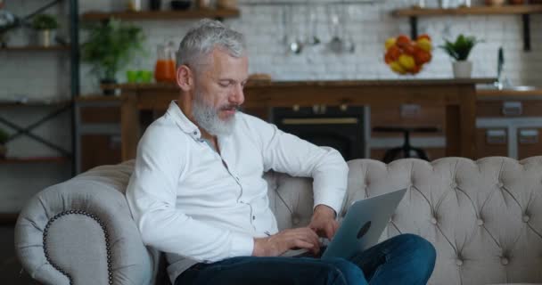 Portret białego, przystojnego, dojrzałego brodatego mężczyzny z siwymi włosami siedzącego na kanapie w salonie w apartamencie i piszącego na laptopie, pracującego zdalnie w domu — Wideo stockowe