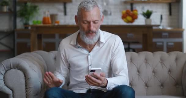 Amazed happy mature businessman receiving message reading good news at living room. Excited overjoyed male winner celebrating success looking at smart phone on sofa at home. Mobile victory concept — Wideo stockowe