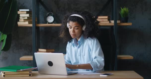 ビデオ会議の呼び出しを介してオンラインチューターの先生とヘッドセットの学習研究を持つ幸せなアフリカの女性学生、ノートパソコンのコンピュータ画面で自宅のオフィスの机を見て座って作る — ストック動画