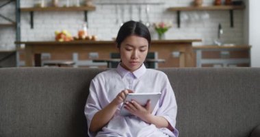 Evdeki dijital bilgisayar tabletini kullanarak rahat koltukta oturan çekici Asyalı genç kadın. Rahat kadın iş kadını internet sörfü bilgisi, evden tek başına çalışıyor.