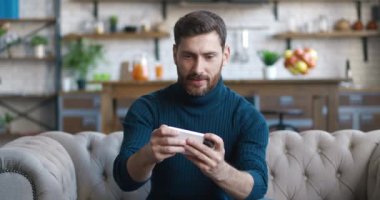 Endişeli genç oyuncu akıllı telefon uygulaması ile çevrimiçi video oyunu oynuyor, evdeki koltukta oturuyor, online oyun uygulamasının kaybından dolayı hayal kırıklığına uğruyor.