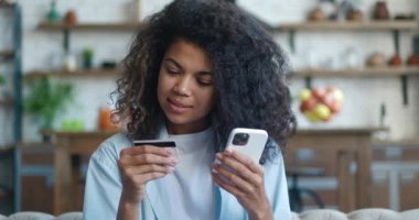 Gülümseyen Afro-Amerikan genç kadın oturma odasında koltukta oturuyor, elinde kredi kartı ve cep telefonu ile online alışveriş yapıyor, dilek tutulan şeyleri alıyor, ev konseptinden alışveriş yapıyor.