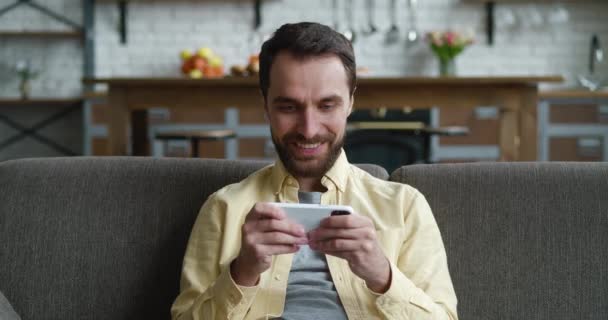 Joven jugando en el teléfono inteligente emocionante aplicación móvil ganar un juego de carreras apretando puño regocijo mientras se sienta en el sofá acogedor en la sala de estar en casa. — Vídeos de Stock