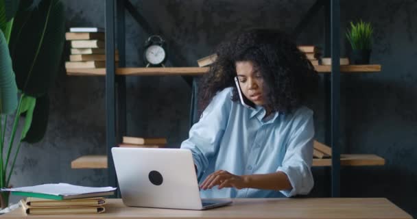 Молодая занятая деловая женщина-профессионал разговаривает по телефону с помощью ноутбука, сидя за столом офиса. Африканская студентка-американка делает мобильный звонок во время работы за компьютером на современном рабочем месте — стоковое видео