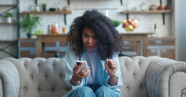 Verontruste jonge Afrikaanse Amerikaanse vrouw reageert negatief op het verliezen van een gokken inzet in een internet casino terwijl zittend op de bank thuis. ongelukkige depressieve jonge vrouw staren naar mobiele telefoon scherm. — Stockvideo