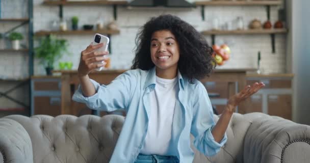 Lachende jonge Afro-Amerikaanse vrouw met krullend haar die videogesprekken voert terwijl ze thuis op een bank zit. Portret van een mooie vrouw die een video chat doet op de achtergrond van de woonkamer — Stockvideo