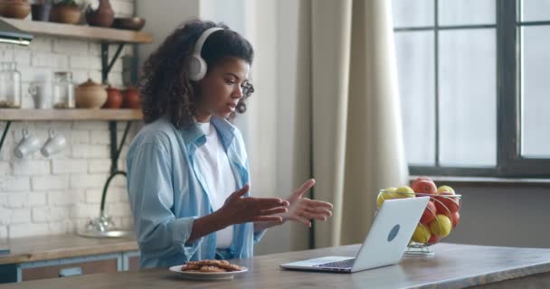 Весела афро-американська молода студентка сидить за кухонним столом на ноутбуці і розмовляє з другом онлайн з бездротовими навушниками з її вух . — стокове відео