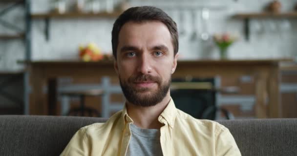 Primer plano retrato del hombre sonriente está descansando mientras está sentado en el sofá en casa. Joven hombre millennial disfruta del estilo de vida mirando a la cámara mientras está sentado en un acogedor sofá en su apartamento. — Vídeo de stock