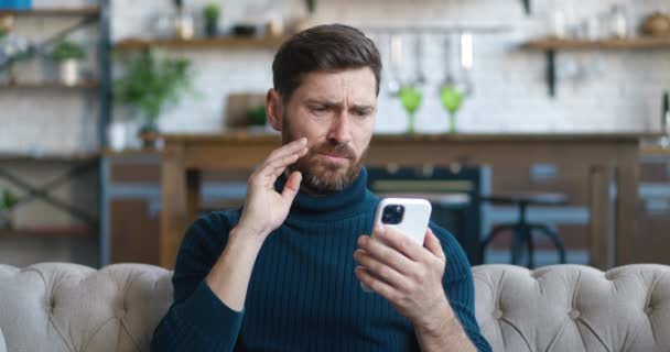 Hombre molesto reaccionar mensaje en el teléfono inteligente se cubre la boca de miedo sentado en el sofá en la sala de estar en casa. Desagradablemente sorprendido y asustado. Hombre se sorprende por lo que vio en la pantalla del teléfono móvil — Vídeos de Stock