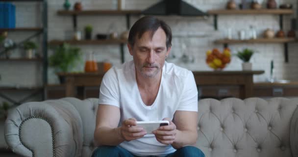 Беспокойный зрелый мужчина геймер играть в видеоигры онлайн с приложением смартфона, сидя на диване дома, разочарован потерей онлайн игры приложения — стоковое видео
