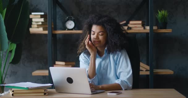 Переживала африканская деловая женщина с вьющимися волосами, работающая на ноутбуке в современном офисе. Скучная женщина-менеджер устала работать за компьютером на своем рабочем месте — стоковое видео