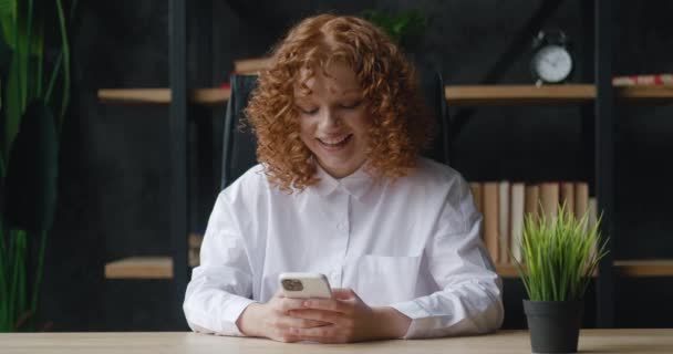 Smiling red-haired young woman holding modern smartphone texting message sitting of office. Young female office worker using modern mobile phone technology app, typing email sitting workplace — Stock Video
