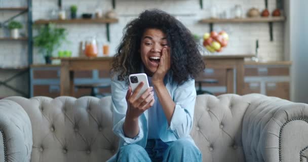 Emocionada mujer afroamericana ganadora celebrando la victoria móvil en la sala de estar, mostrando sí gesto. Mujer encantada con el regalo ganador de teléfonos inteligentes o premio en la aplicación de redes sociales, en el sofá en casa. — Vídeo de stock