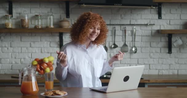 Joven mujer pelirroja con el pelo rizado utilizando el ordenador portátil para la conexión de vídeo remoto, participa en reuniones virtuales, hablar y discutir en línea, participar en videollamadas, sentarse en la cocina en casa — Vídeos de Stock