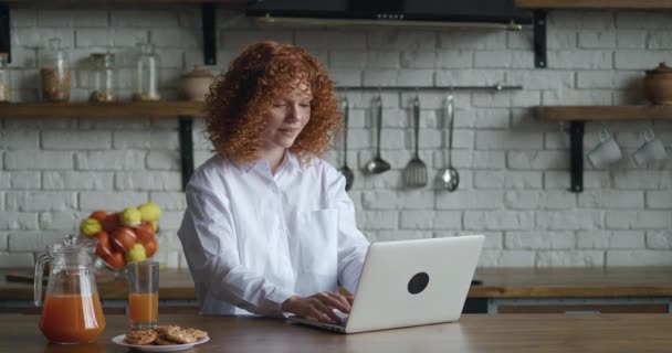 Heyecanlı kızıl saçlı genç iş kadını laptopuyla masada oturuyor ve ofis mutfağında çalışıyor. Harika haberler alıyor ve çok mutlu. Evet işareti gösteriliyor — Stok video