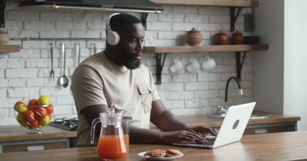 부엌에 랩탑있는 아프리카 우승자. 헤드폰을 끼고 있는 행복 한 남자 가 집 부엌의 식탁에 앉아 있다가 복권에 당첨된 후 흥분 한 채 노트북을 보고 있습니다. — 비디오