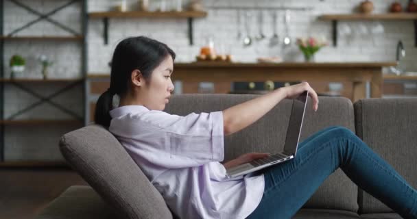 Müde junge asiatische freie Frau beenden ihre Arbeit am Computer und ruhen sich auf der Couch aus. Weibchen schließt Laptop, hebt Arme, dehnt Muskeln, hält Fristen ein, beendet Arbeitstag zu Hause. — Stockvideo