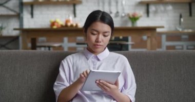 Evdeki dijital bilgisayar tabletini kullanarak rahat koltukta oturan çekici Asyalı genç kadın. Rahat kadın iş kadını internet sörfü bilgisi, evden tek başına çalışıyor.
