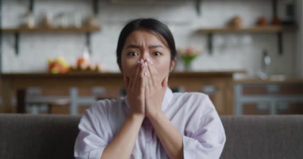 Portret wystraszonej Azjatki, reakcja na złe wieści, siedzenie na kanapie w domu. Zszokowana azjatycka kobieta patrząca w kamerę ze strachu przed strasznymi wiadomościami — Wideo stockowe