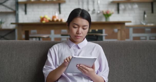 Attraktive junge Asiatin, die auf einem bequemen Sofa sitzt und zu Hause ein digitales Computer-Tablet benutzt. Entspannte Geschäftsfrau surft im Internet und arbeitet allein von zu Hause aus — Stockvideo